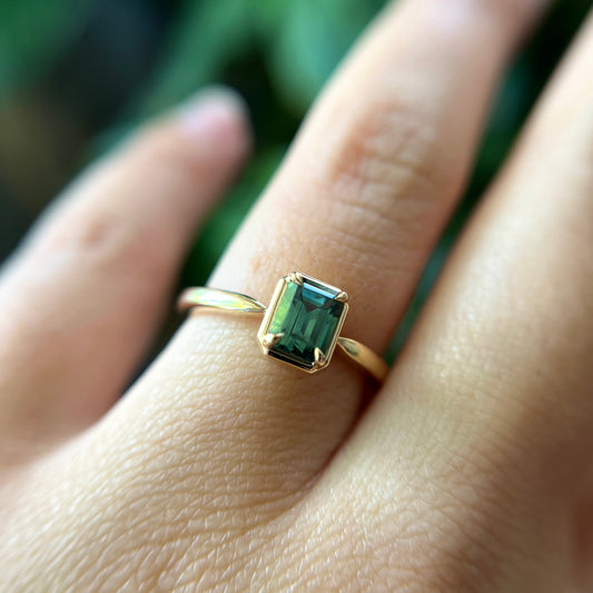Bague de fiançailles solitaire en saphir vert forêt taille émeraude de 1,05ct de Madagascar en or jaune 14k, portée au doigt montrant la couleur de la pierre et le serti en griffes à cadre en or