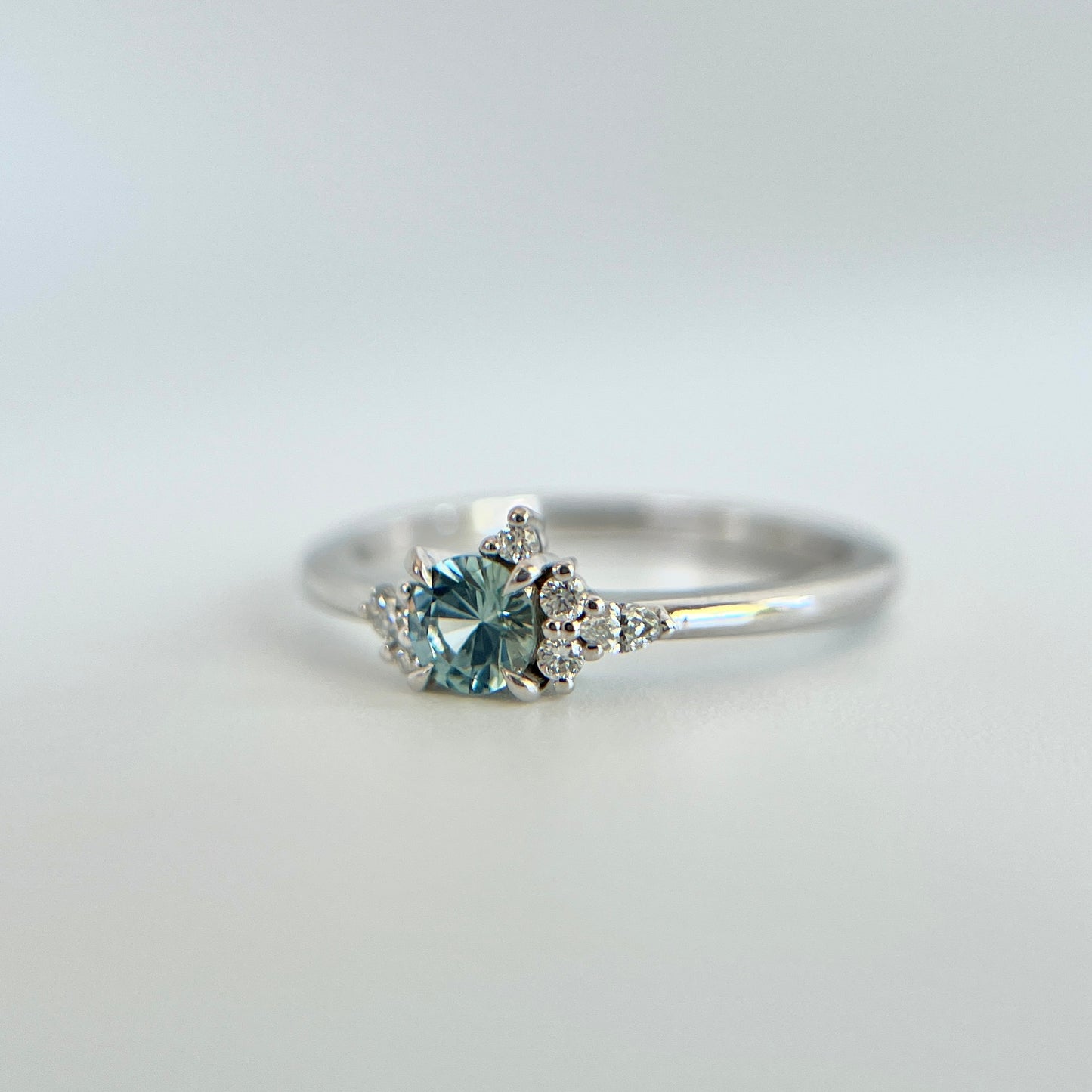 Bague de fiançailles en saphir bleu rond de 0,35ct et diamants naturels en or blanc 14k, vue en trois-quarts montrant la couleur de la pierre et le détail du cluster asymétrique