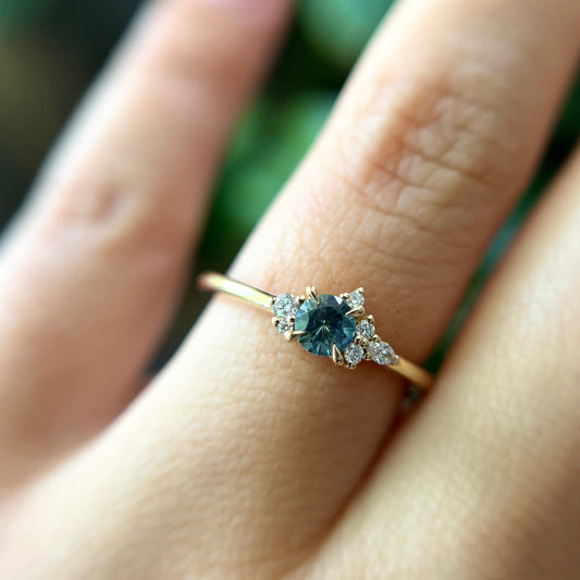 Bague de fiançailles en saphir sarcelle rond de 0,38ct et diamants naturels en or jaune 14k, portée au doigt montrant la couleur de la pierre et le cluster asymétrique