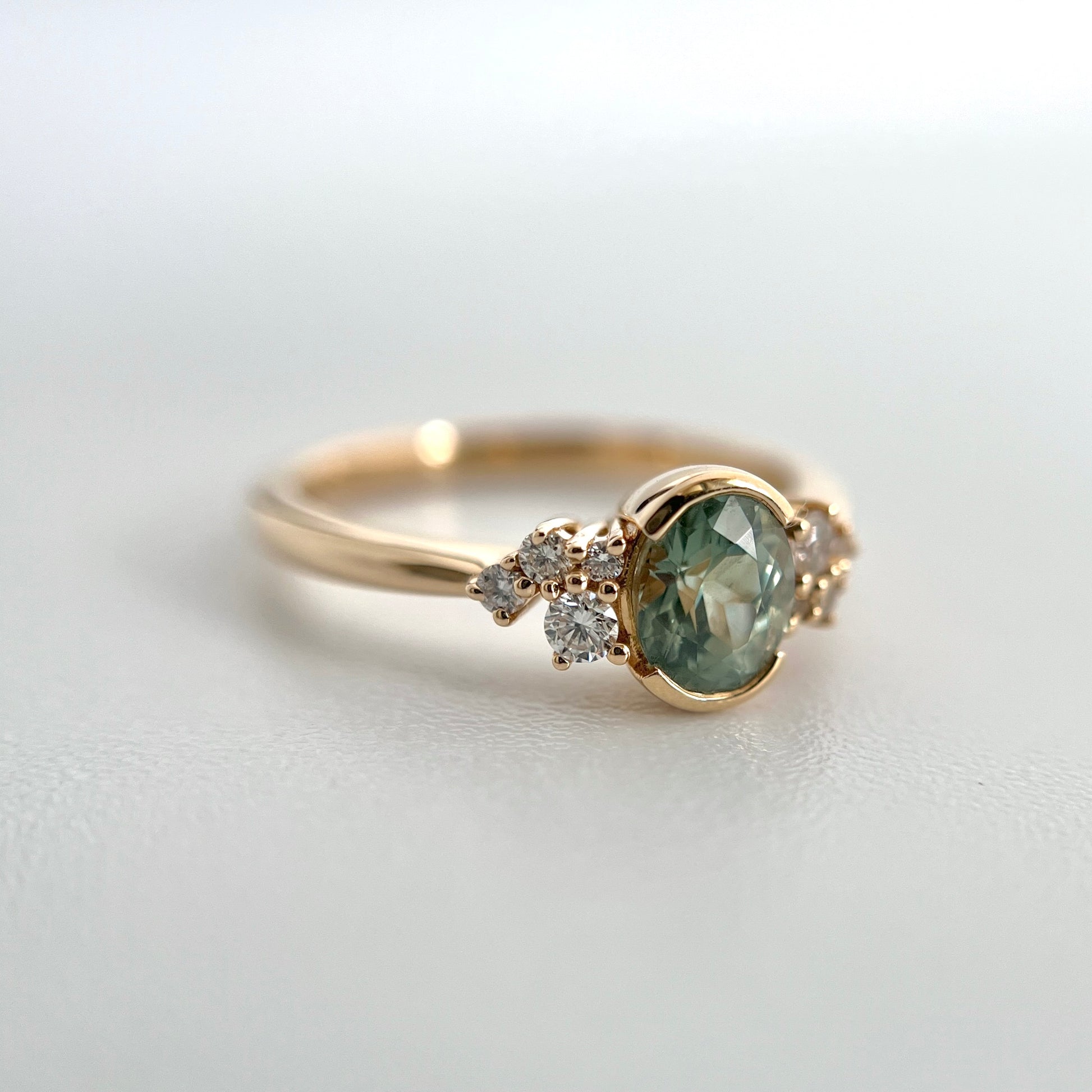 3/4 view of an engagement ring, half-bezel set with a light green-teal sapphire from madagascar, and a cluster of 4 diamonds on each side. This photo shows the diamond and cluster details. 