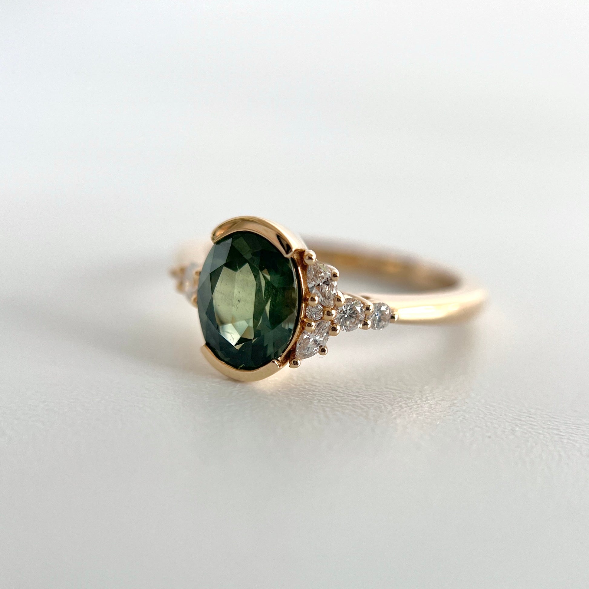 Oval green half-bezel set sapphire and natural diamond ring. This view showcases the marquise and round diamond accents on one side of the ring.