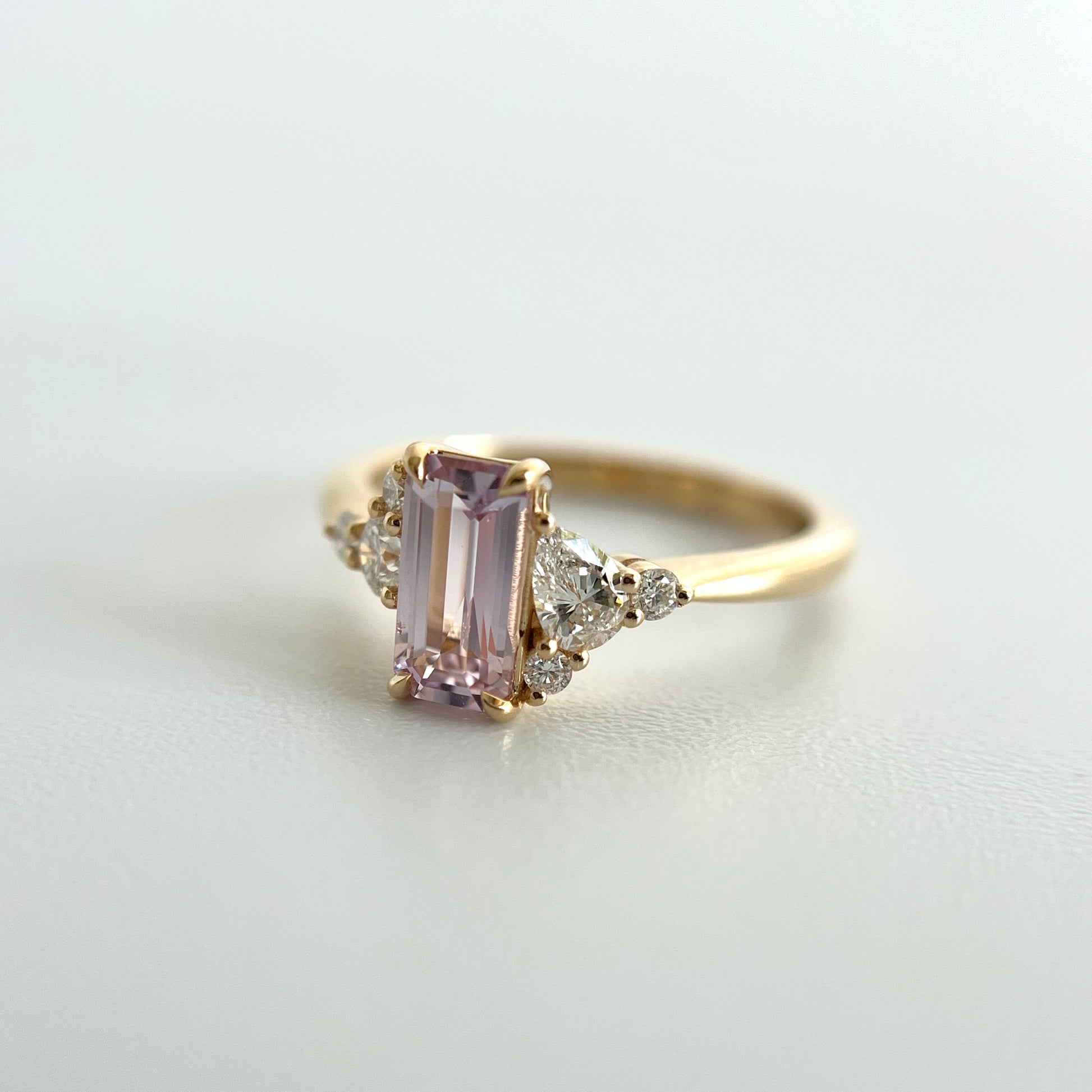 Angled view of a 14k yellow gold ring, eagle claw set with a light pink emerald-cut sapphire, and natural diamond accents. This photo puts an emphasis on the larger pear shape and two smaller round diamond accents on the right side of the ring. 