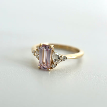 Angled view of a 14k yellow gold ring, eagle claw set with a light pink emerald-cut sapphire, and natural diamond accents. This photo puts an emphasis on the larger pear shape and two smaller round diamond accents on the right side of the ring. 