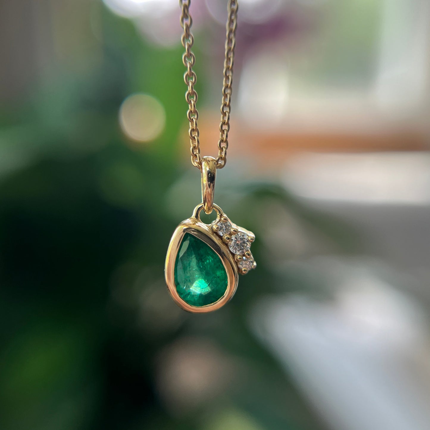 A gold pendant necklace featuring a pear-shaped green emerald and a small cluster of clear stones, hanging against a softly blurred green and white background.