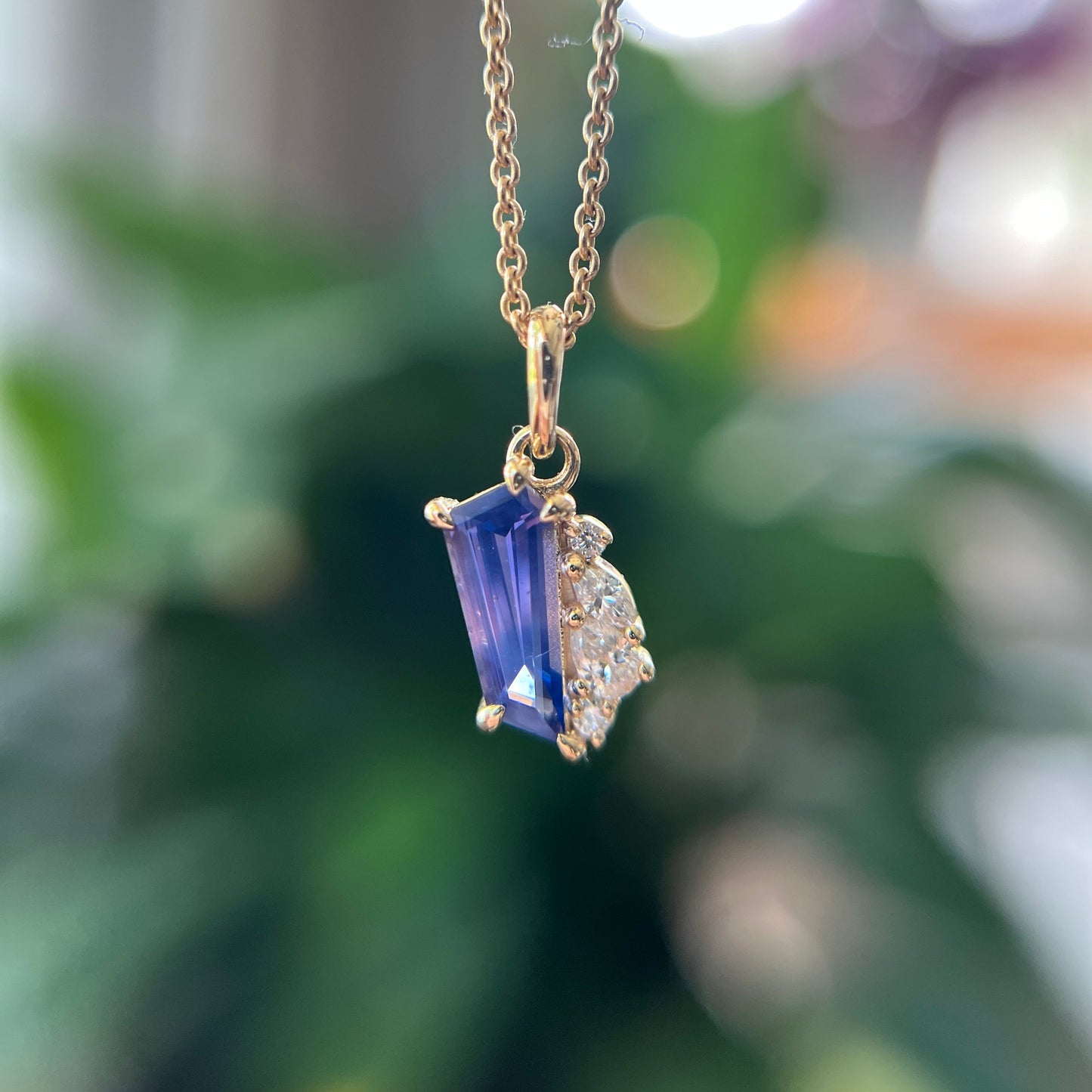 A gold pendant necklace featuring a freeform blue to pink sapphire and a small cluster of natural diamonds, shown hanging against a blurred green background.