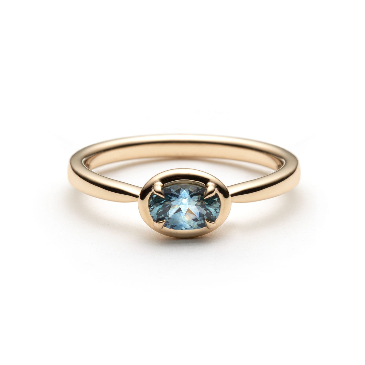 Denim blue oval montana sapphire set horizontally between 4 claws, in a frame bezel-like setting. The solitaire is shown on top of a white background. 