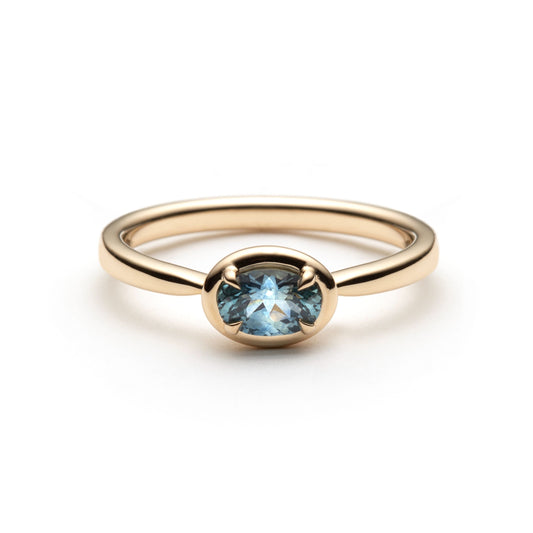 Denim blue oval montana sapphire set horizontally between 4 claws, in a frame bezel-like setting. The solitaire is shown on top of a white background. 