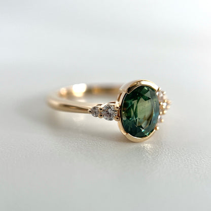 14k yellow gold ring, set with an oval green sapphire in a half-bezel and two round brilliant cut natural diamonds on one side. 