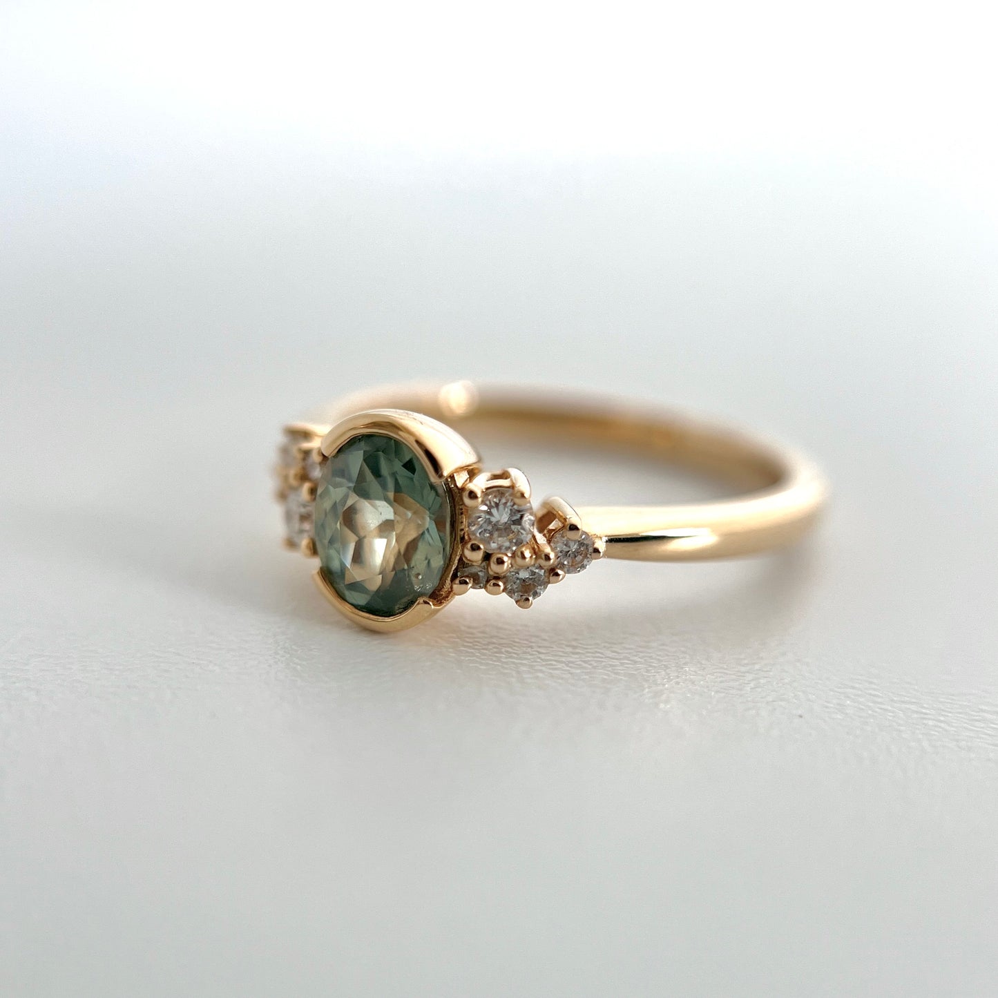 A yellow gold ring featuring an oval green-teal sapphire in a half bezel setting, accented by small diamonds on either side, photographed on a light gray surface.
