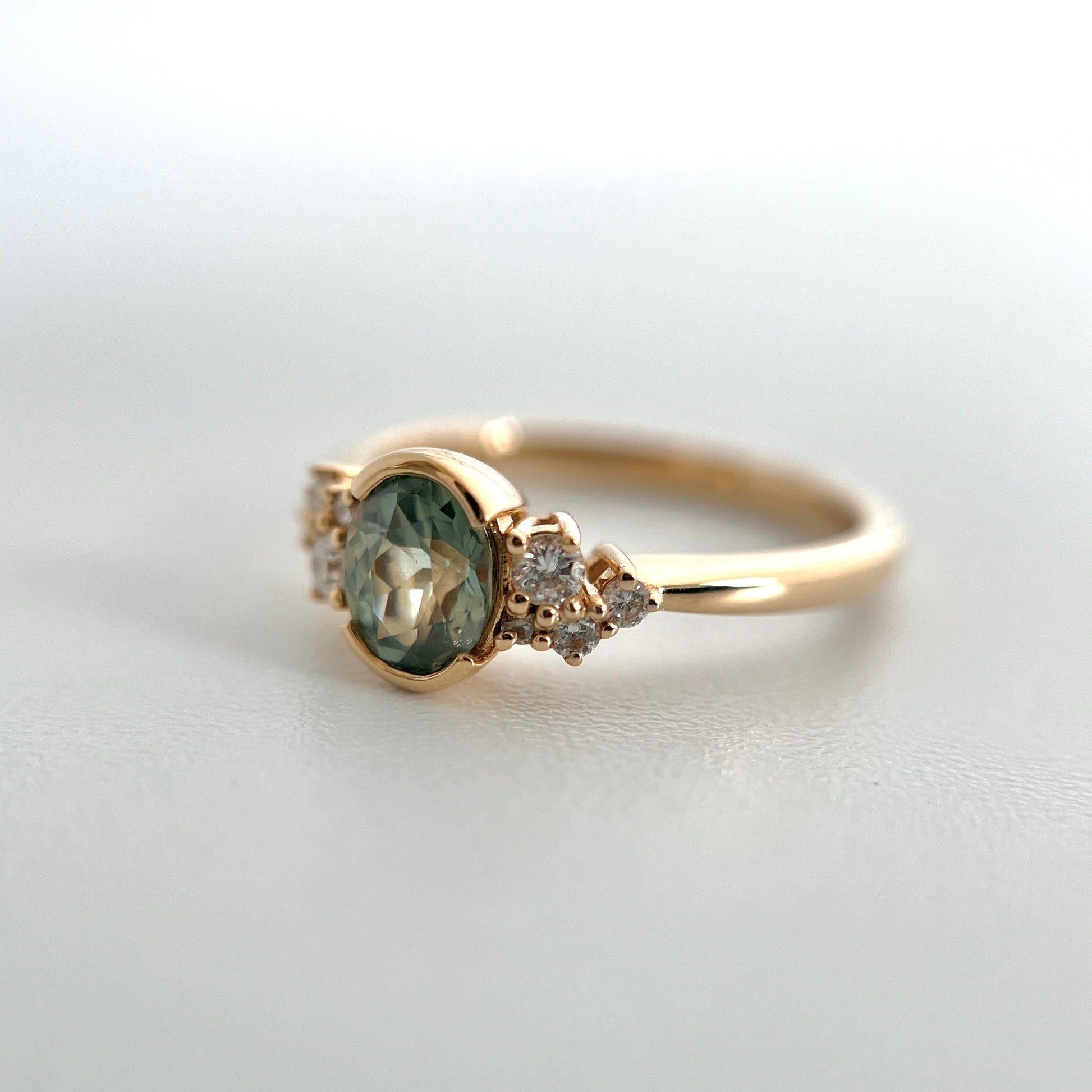 A yellow gold ring featuring an oval green-teal sapphire in a half bezel setting, accented by small diamonds on either side, photographed on a light gray surface.