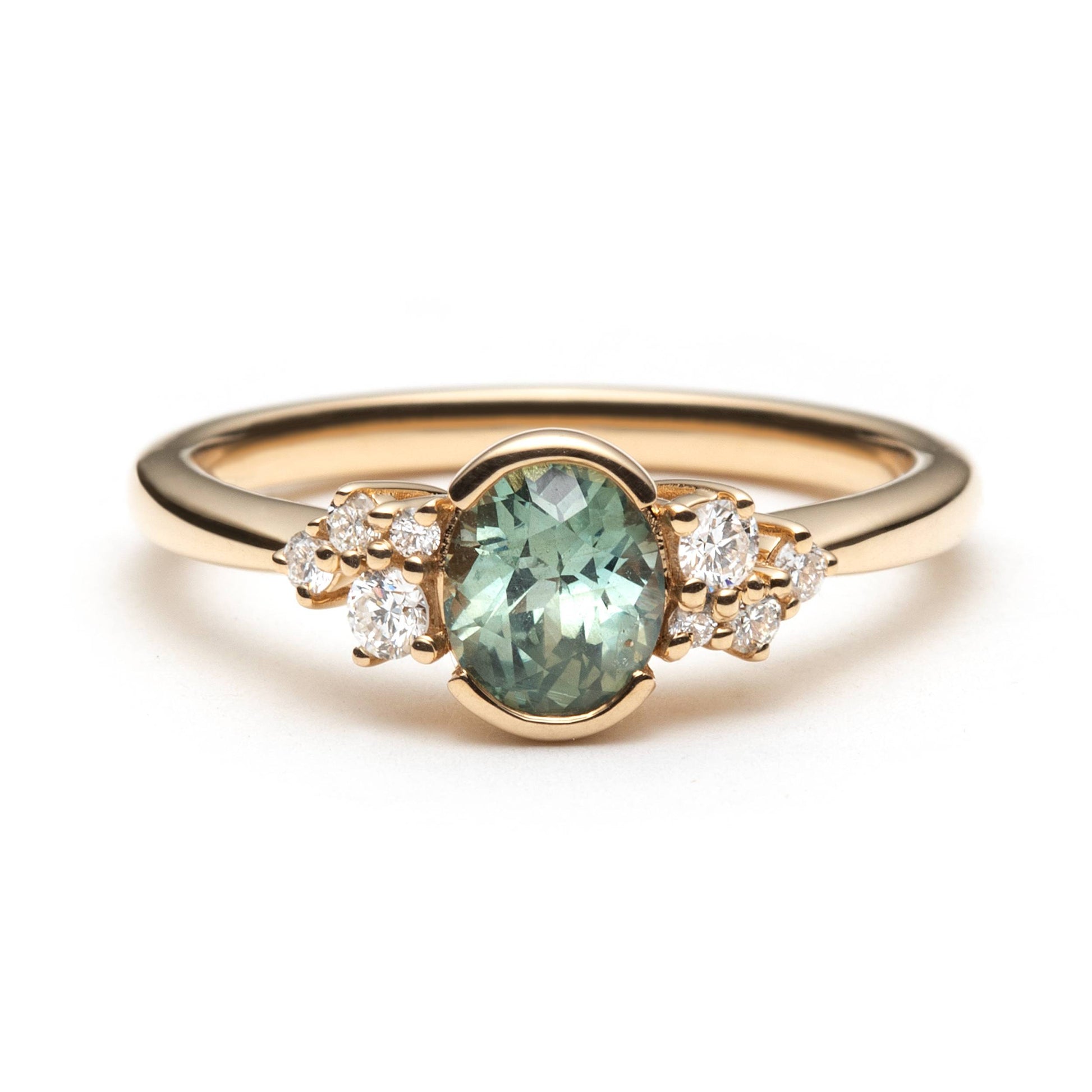 Oval teal sapphire bezel set in 14k yellow gold, and accented by two delicate diamond clusters. The ring is on a white background. 