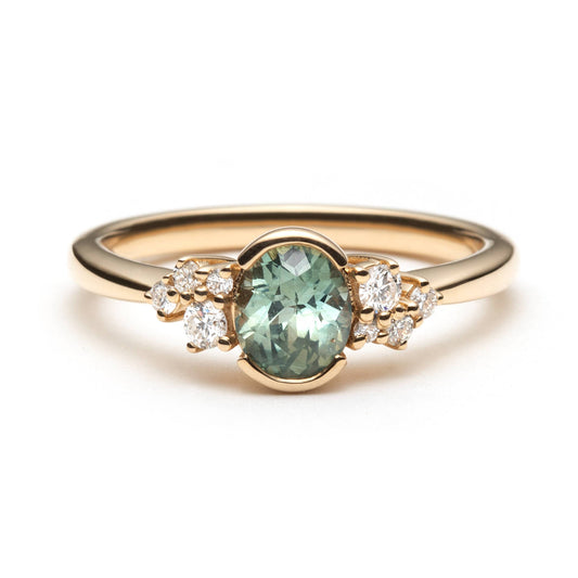 Oval teal sapphire bezel set in 14k yellow gold, and accented by two delicate diamond clusters. The ring is on a white background. 
