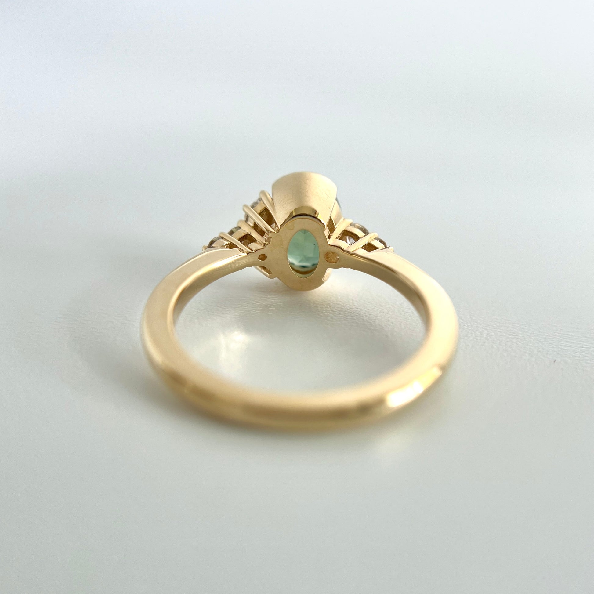 Under side view of a ring set with an oval green sapphire in the middle, with a half-bezel setting, and detailed view of the side open gallery settings for the small diamonds. 