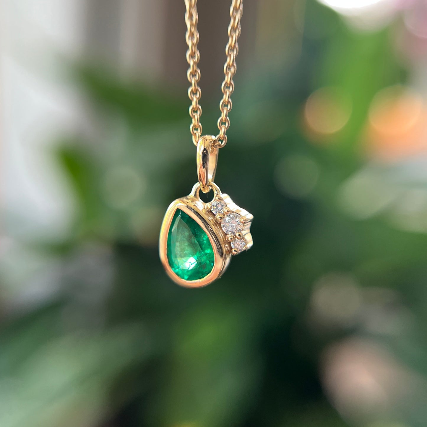 A gold pendant necklace featuring a teardrop-shaped green emerald and a small cluster of clear stones, shown hanging against a blurred green background.