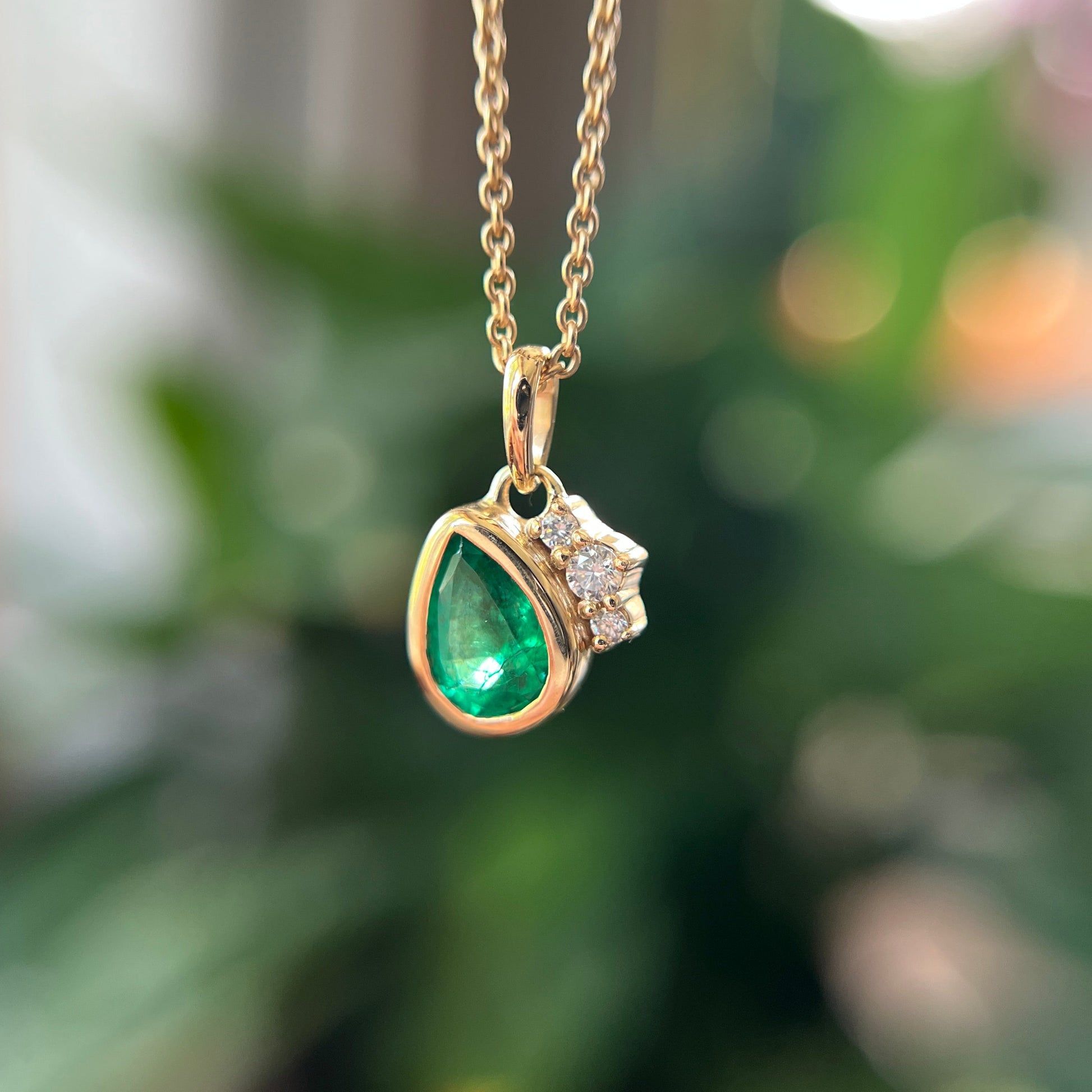 A gold pendant necklace featuring a teardrop-shaped green emerald and a small cluster of clear stones, shown hanging against a blurred green background.
