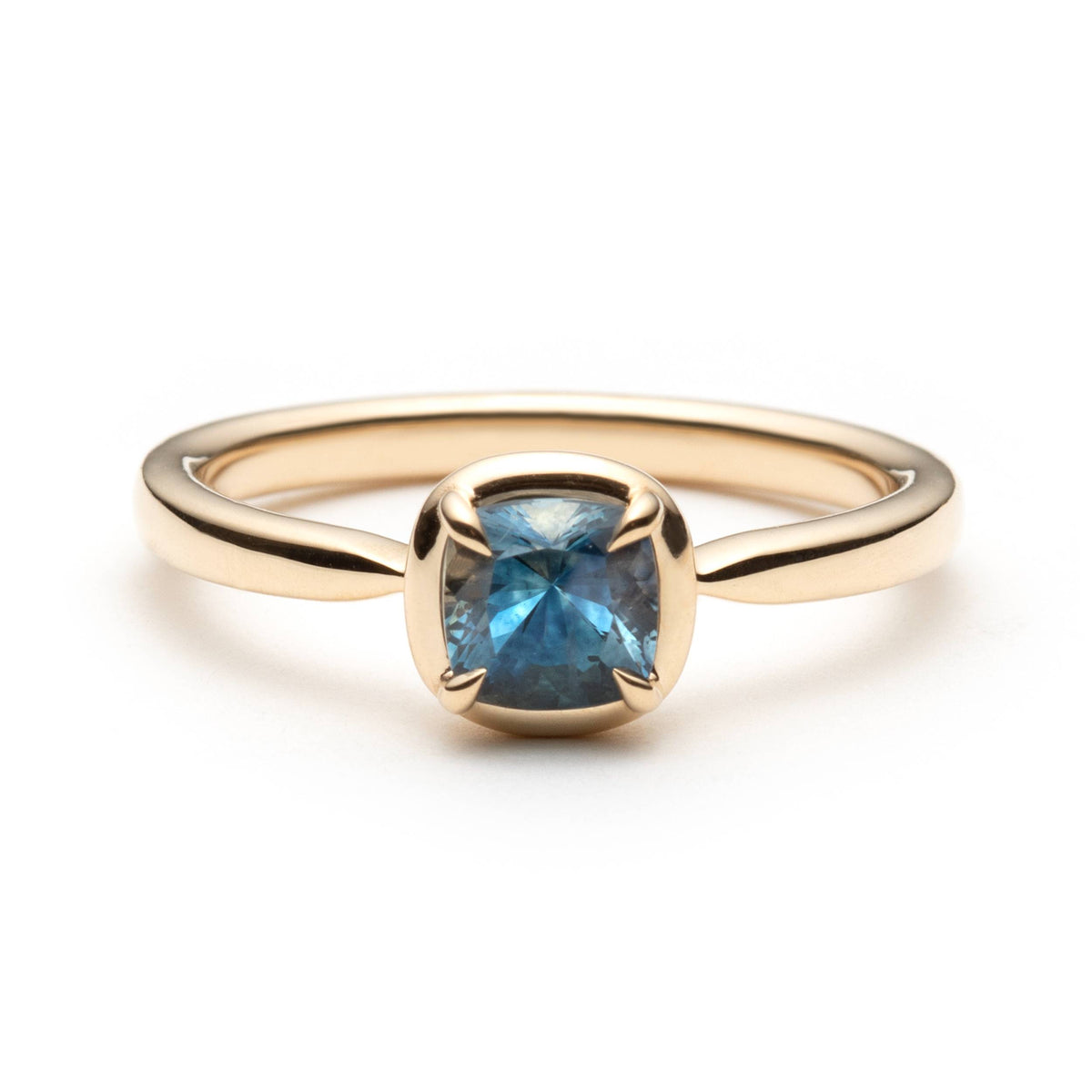 Gold solitaire ring with a denim blue cushion-cut eagle-claw set sapphire, with a gold border, on a white background