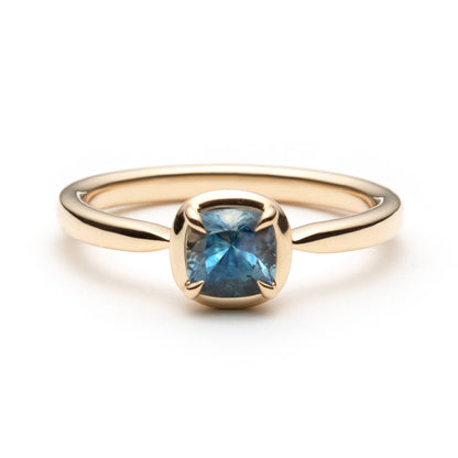 Gold solitaire ring with a denim blue cushion-cut eagle-claw set sapphire, with a gold border, on a white background