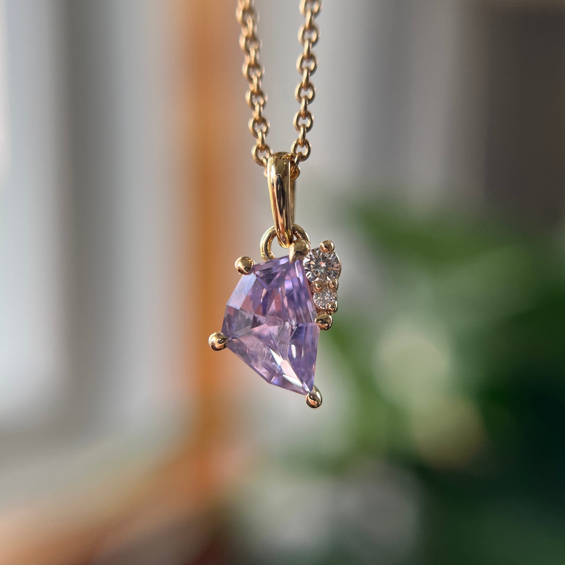 A gold pendant necklace featuring a triangular light purple gemstone and a small cluster of clear stones, hanging from a gold chain against a softly blurred indoor background.