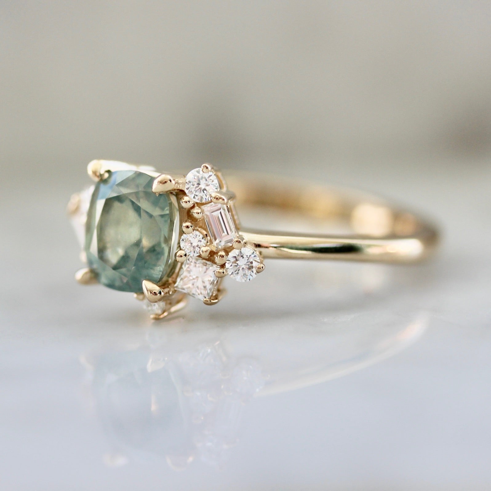 1.47ct unheated cushion cut green Montana sapphire and diamond cluster ring in 14k yellow gold, angled view showing stone colour and asymmetric diamond setting detail