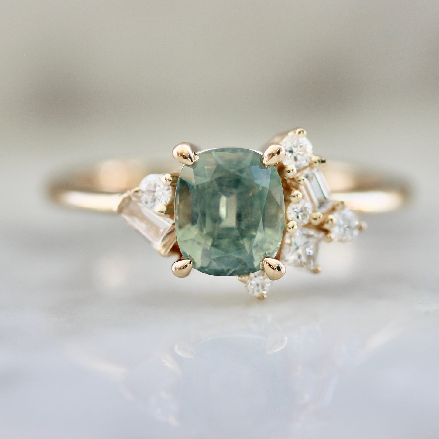 1.47ct unheated cushion cut green Montana sapphire and diamond cluster ring in 14k yellow gold, close-up view showing stone colour and asymmetric baguette and round diamond setting