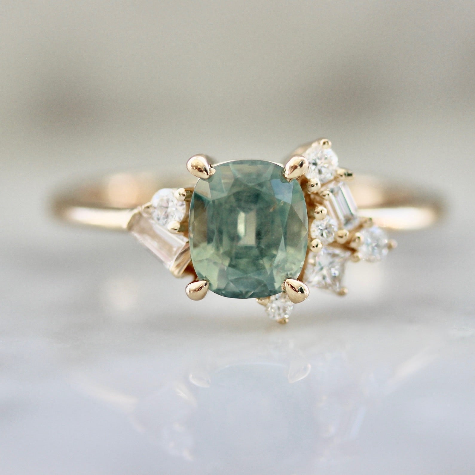 1.47ct unheated cushion cut green Montana sapphire and diamond cluster ring in 14k yellow gold, close-up view showing stone colour and asymmetric baguette and round diamond setting