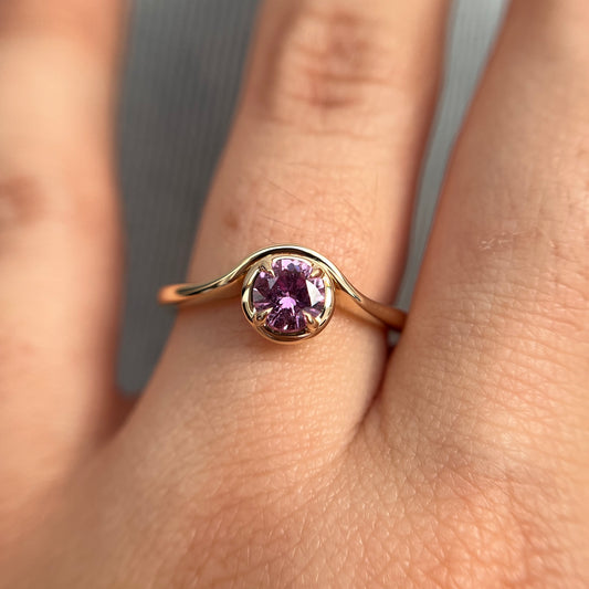 Pink sapphire solitaire ring in 14k yellow gold, worn on hand showing stone colour and wave setting