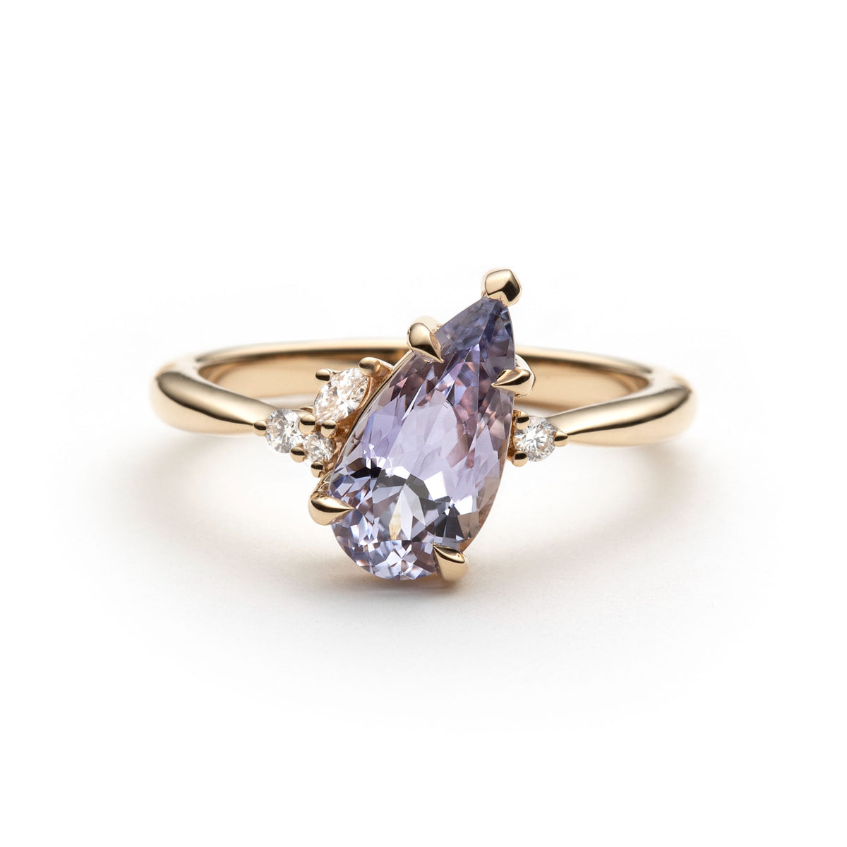 Light purple pear shaped sapphire is set tilted at a 45 degree angle, and surrounded bt 4 asymmetrically placed diamonds. The ring is made in yellow gold, and is shown in front of a white background. 
