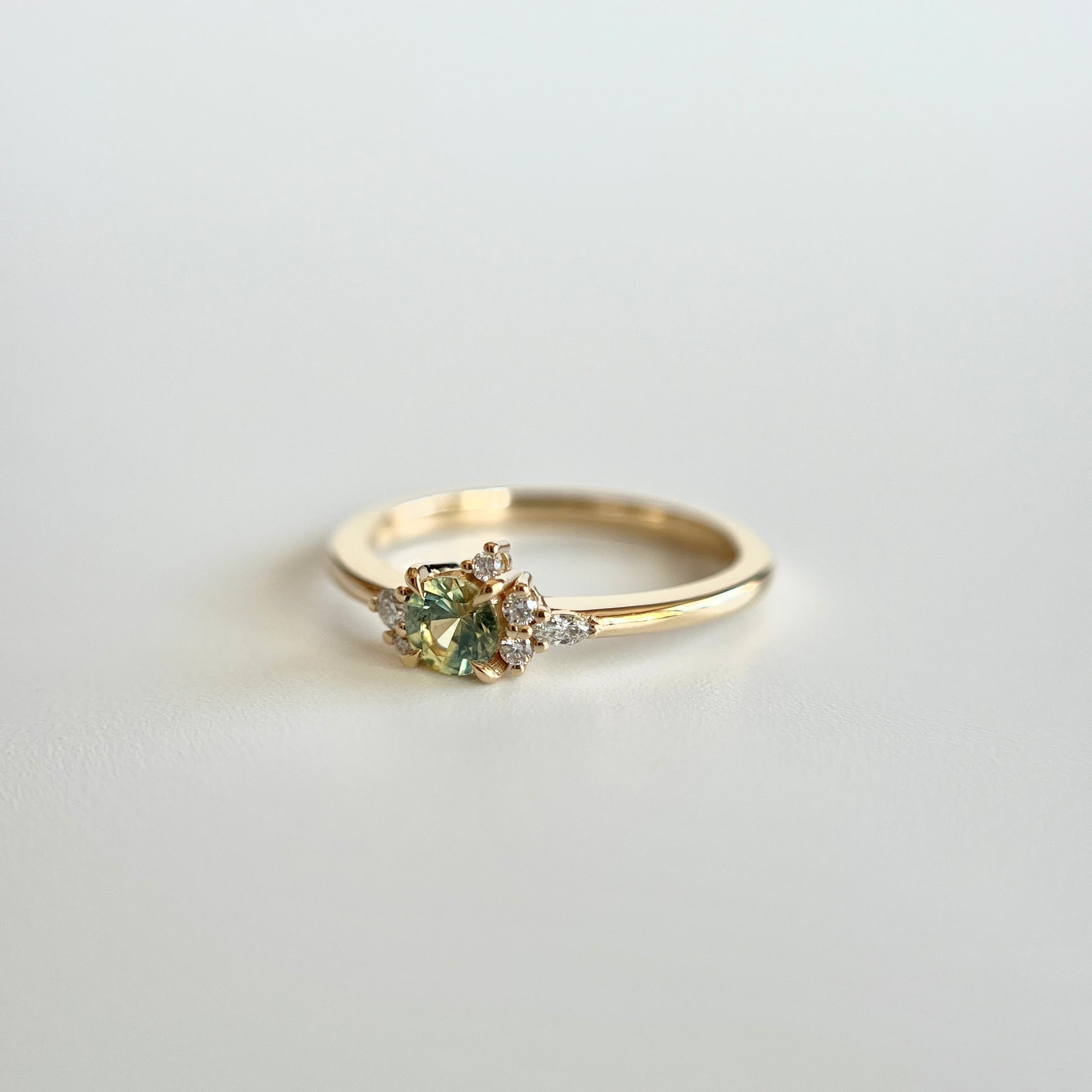 Bague de fiançailles en saphir bicolore rond de 0,36ct passant du jaune au bleu et diamants naturels en or jaune 14k, vue en trois-quarts montrant le changement de couleur de la pierre et le détail du cluster asymétrique