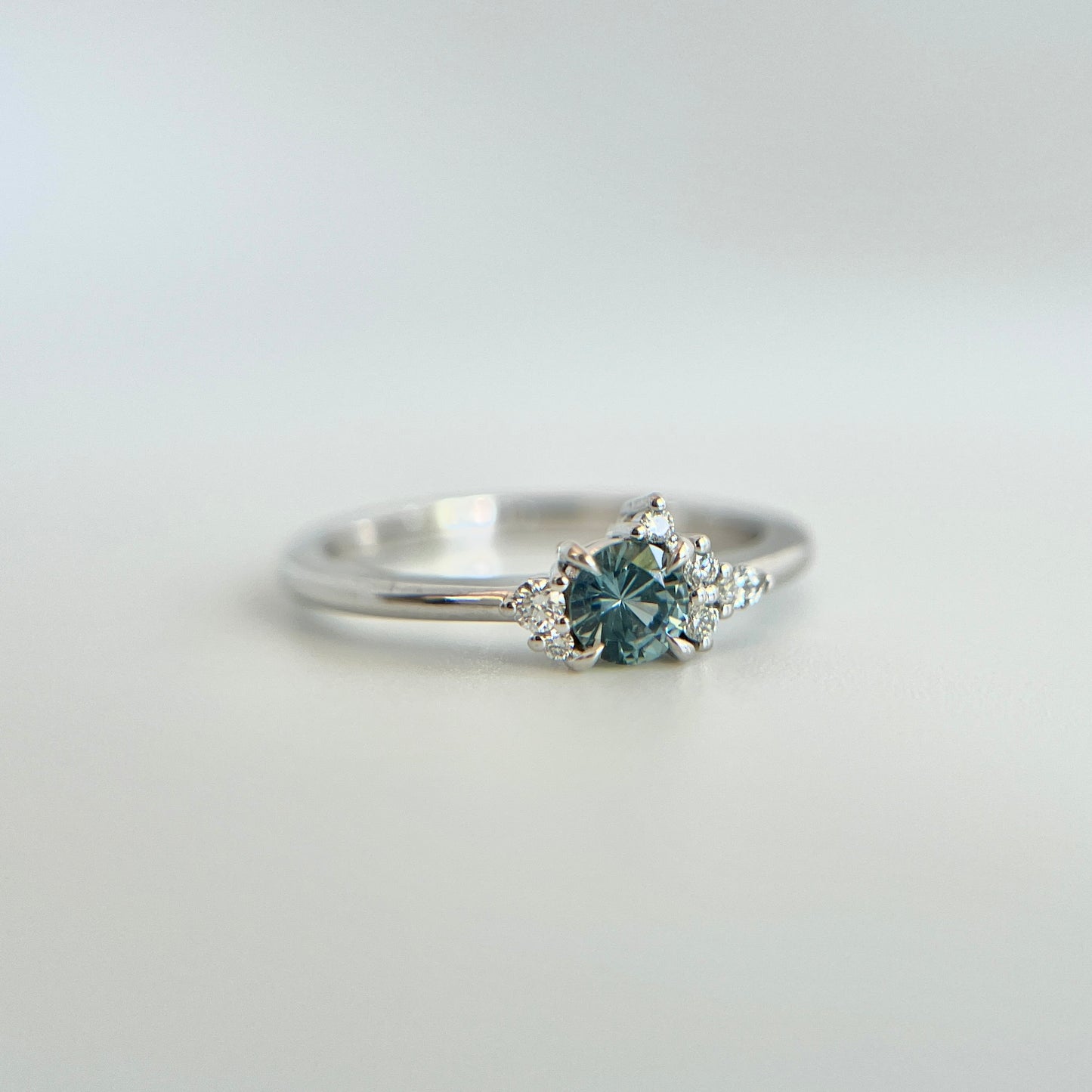 Bague de fiançailles en saphir bleu rond de 0,35ct et diamants naturels en or blanc 14k, vue en angle montrant le cluster asymétrique et le détail du corps de bague