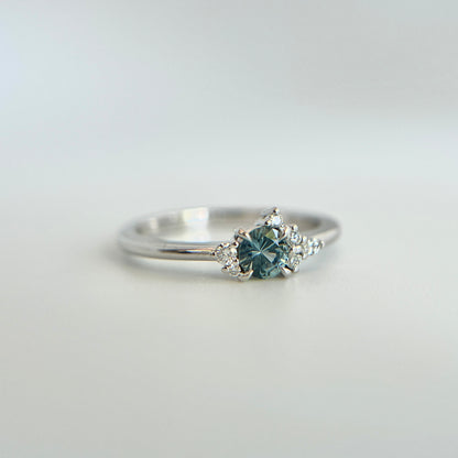 Bague de fiançailles en saphir bleu rond de 0,35ct et diamants naturels en or blanc 14k, vue en angle montrant le cluster asymétrique et le détail du corps de bague