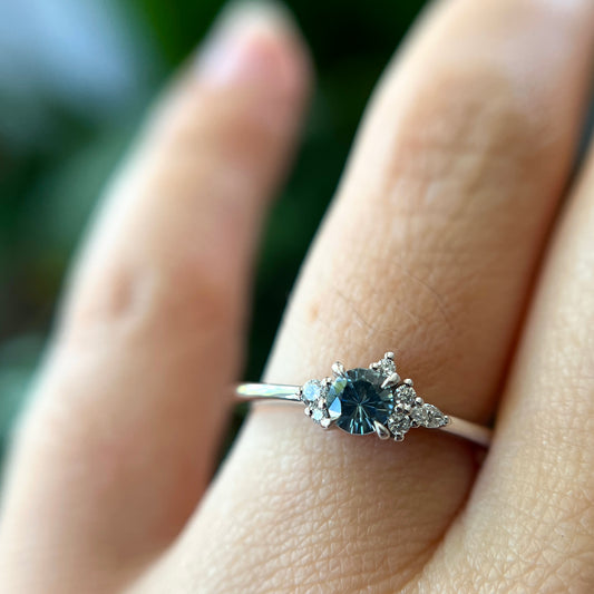 Bague de fiançailles en saphir bleu rond de 0,35ct et diamants naturels en or blanc 14k, portée au doigt montrant la couleur de la pierre et le cluster asymétrique