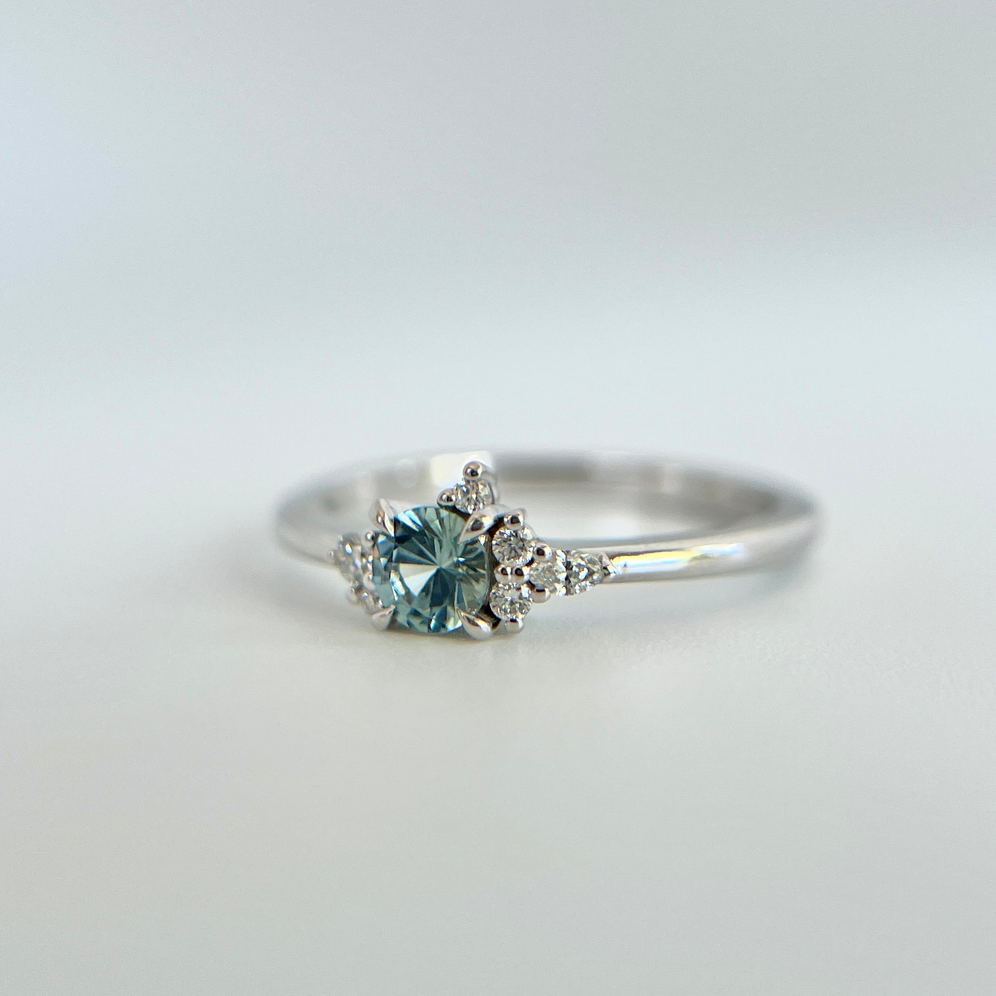 Bague de fiançailles en saphir bleu rond de 0,35ct et diamants naturels en or blanc 14k, vue en trois-quarts montrant la couleur de la pierre et le détail du cluster asymétrique