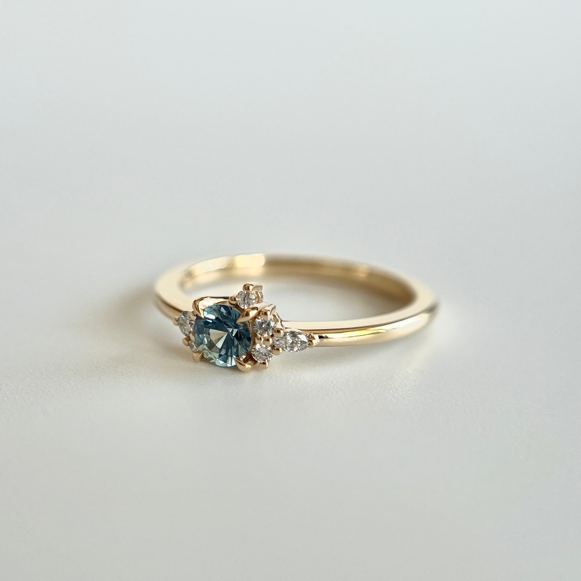 Bague de fiançailles en saphir bleu-sarcelle rond de 0,39ct et diamants naturels en or jaune 14k, vue en angle montrant le cluster asymétrique et le détail du corps de bague