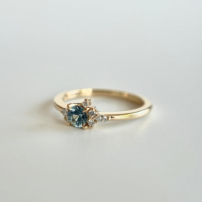 Bague de fiançailles en saphir bleu-sarcelle rond de 0,39ct et diamants naturels en or jaune 14k, vue en angle montrant le cluster asymétrique et le détail du corps de bague