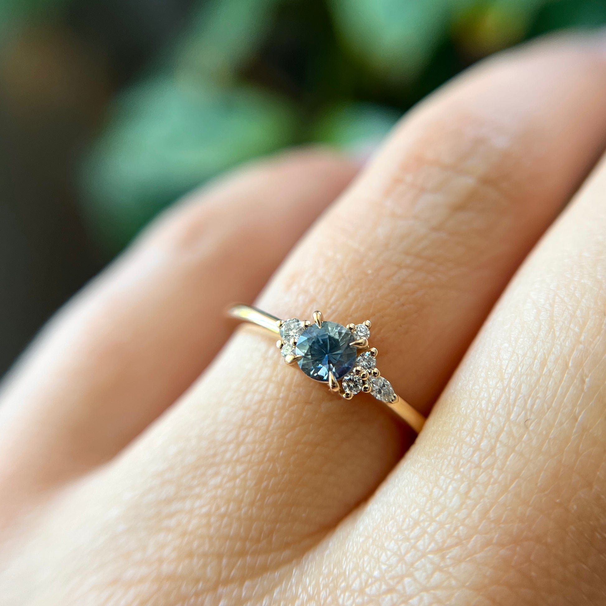 Bague de fiançailles en saphir bleu-sarcelle rond de 0,39ct et diamants naturels en or jaune 14k, portée au doigt montrant la couleur de la pierre et le cluster asymétrique