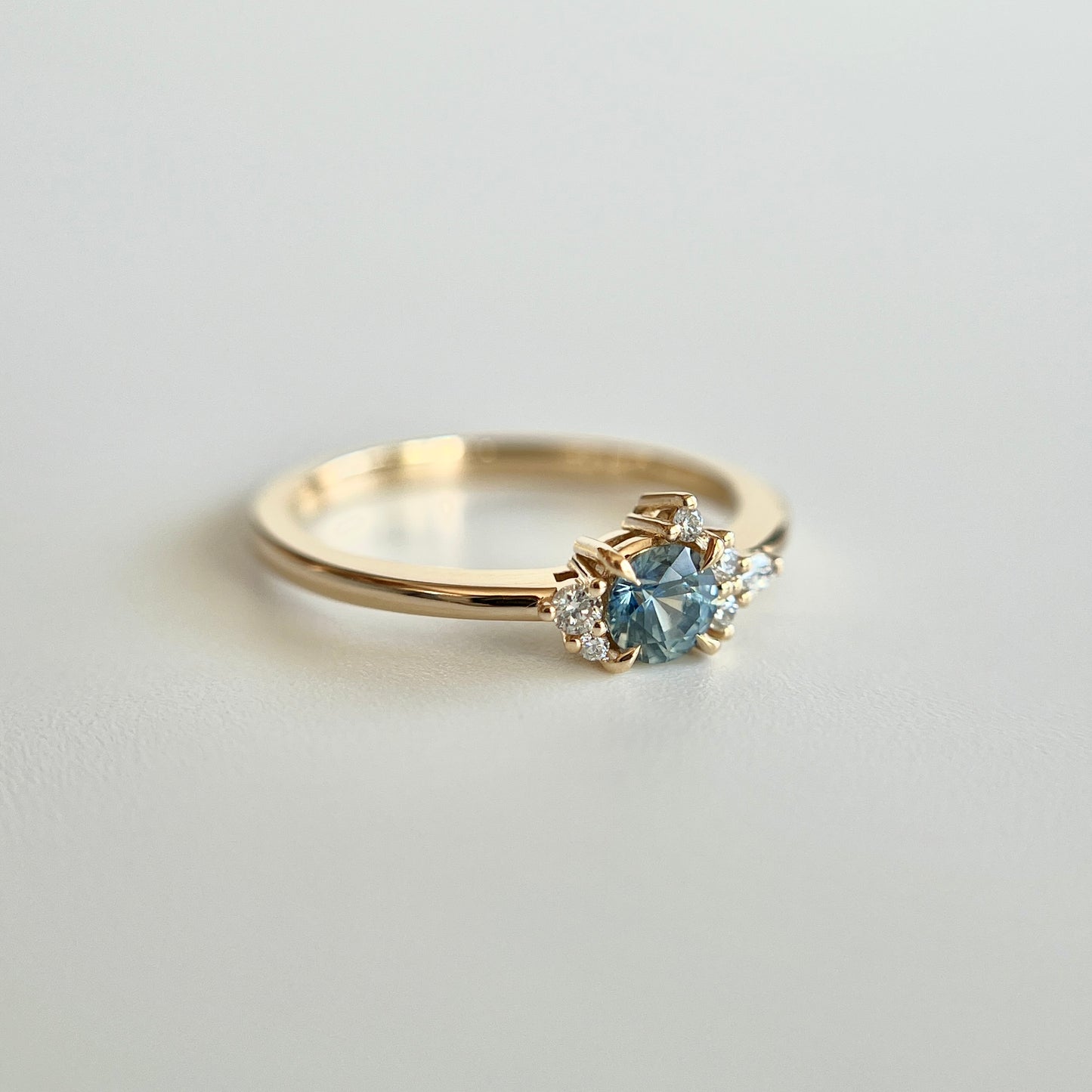 Bague de fiançailles en saphir bleu-sarcelle rond de 0,39ct et diamants naturels en or jaune 14k, vue en trois-quarts montrant la couleur de la pierre et le détail du cluster asymétrique