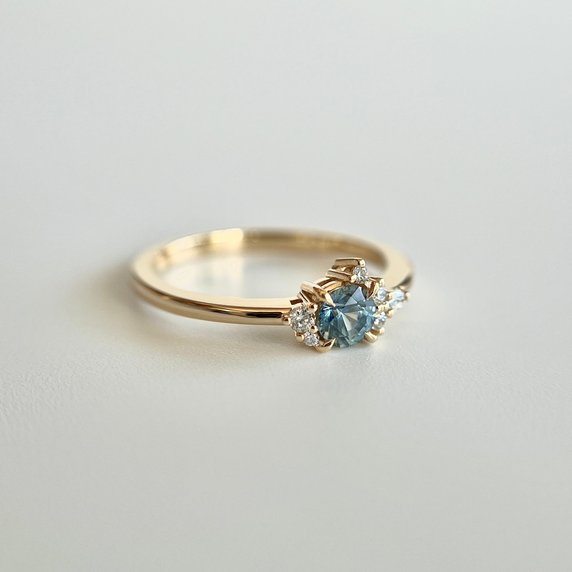 Bague de fiançailles en saphir bleu-sarcelle rond de 0,39ct et diamants naturels en or jaune 14k, vue en trois-quarts montrant la couleur de la pierre et le détail du cluster asymétrique