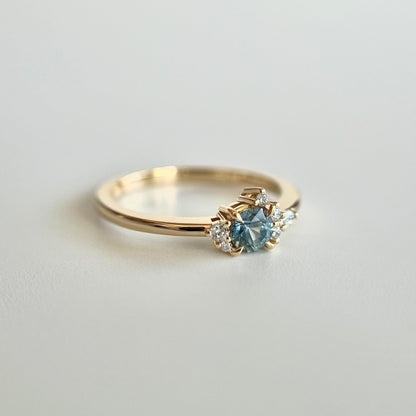 Bague de fiançailles en saphir bleu-sarcelle rond de 0,39ct et diamants naturels en or jaune 14k, vue en trois-quarts montrant la couleur de la pierre et le détail du cluster asymétrique