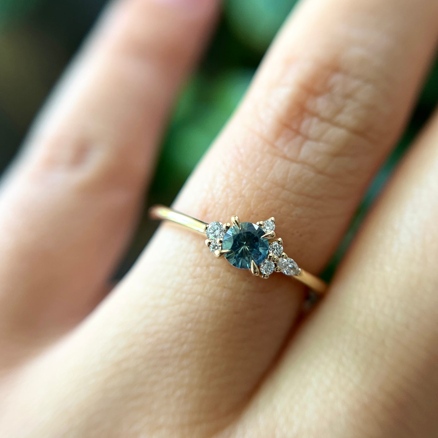 Bague de fiançailles en saphir sarcelle rond de 0,38ct et diamants naturels en or jaune 14k, portée au doigt montrant la couleur de la pierre et le cluster asymétrique