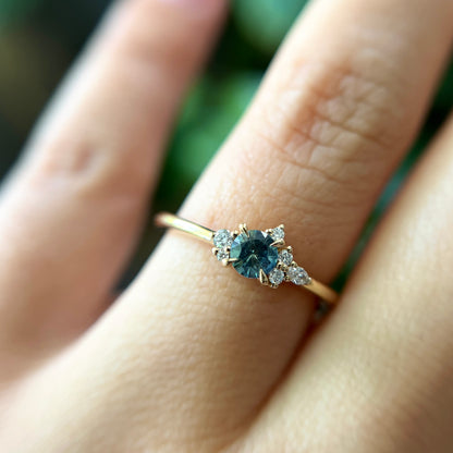 Bague de fiançailles en saphir sarcelle rond de 0,38ct et diamants naturels en or jaune 14k, portée au doigt montrant la couleur de la pierre et le cluster asymétrique