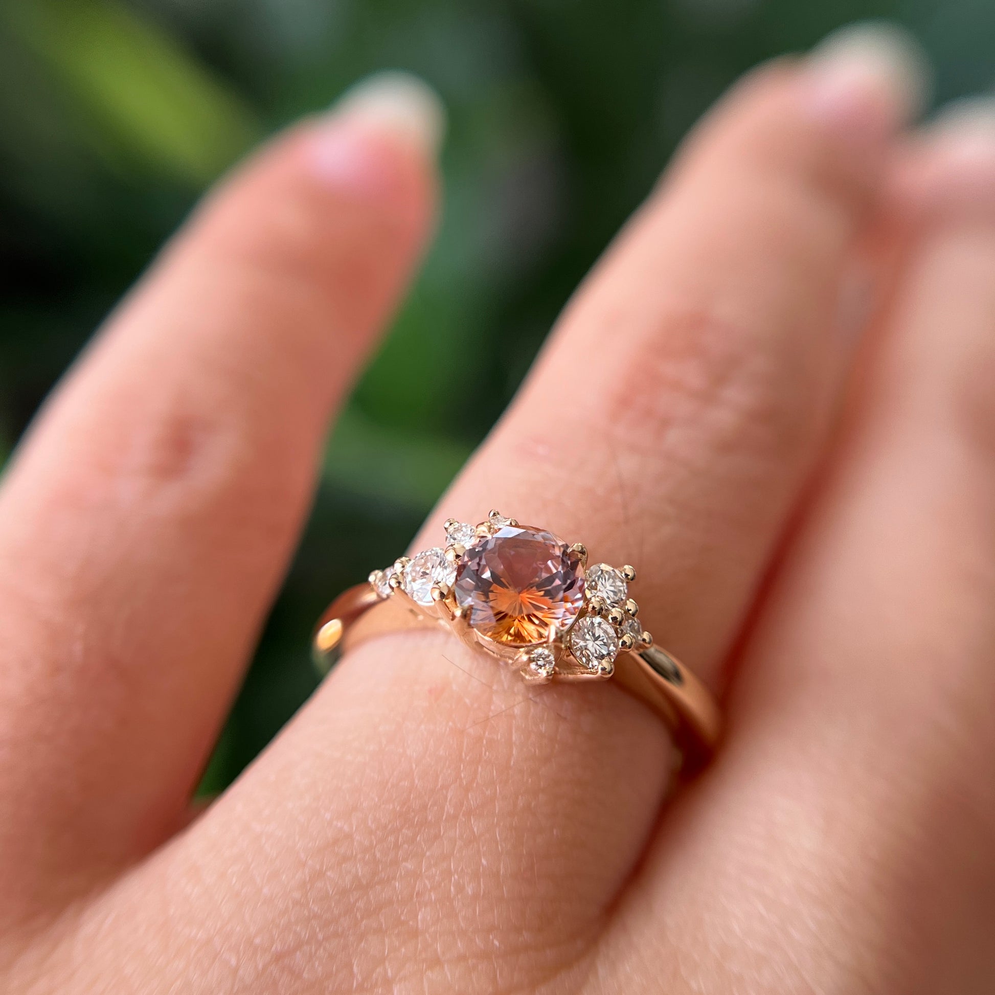 Gros plan de la bague de fiançailles en saphir Montana orange-rose et diamants en or jaune 14k, portée au doigt montrant le changement de couleur