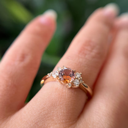 Gros plan de la bague de fiançailles en saphir Montana orange-rose et diamants en or jaune 14k, portée au doigt montrant le changement de couleur