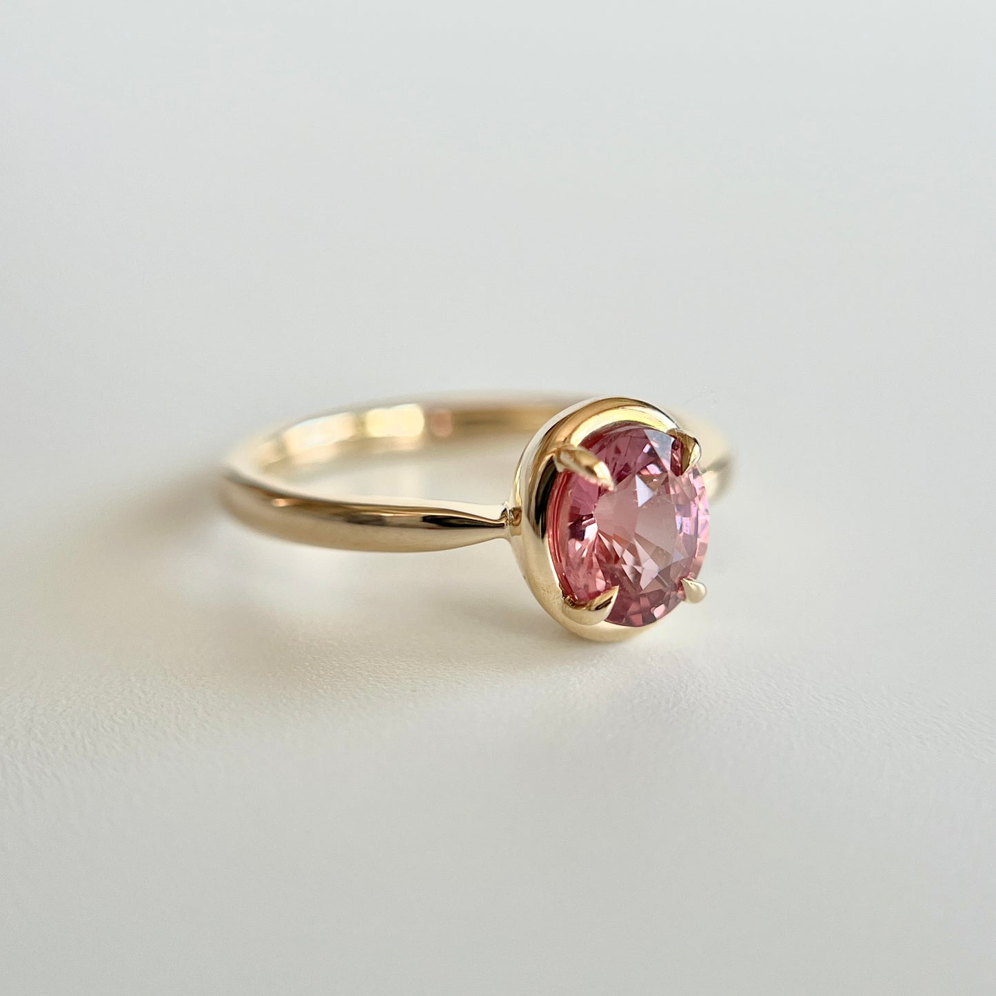 Bague de fiançailles solitaire en saphir ovale rose-orangé de 1,20ct en or jaune 14k, vue en angle montrant le détail du serti en griffes à cadre en or