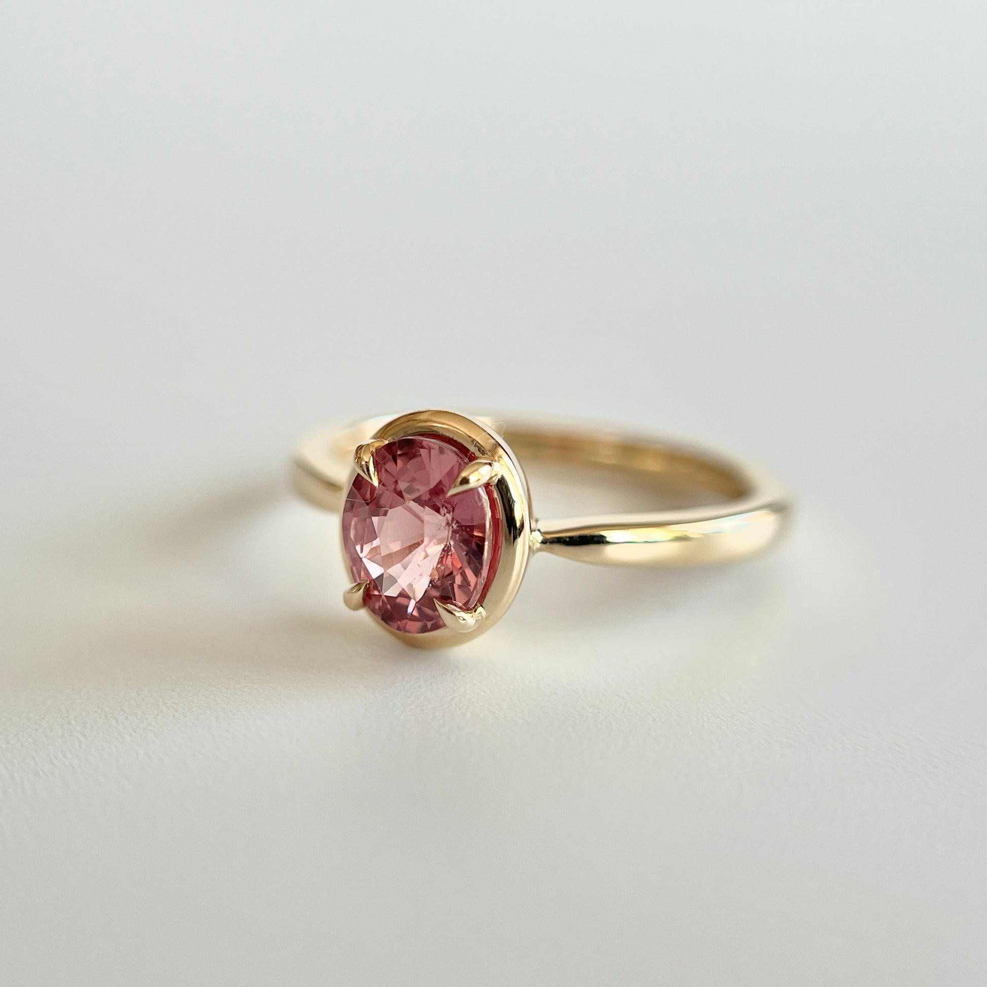 Bague de fiançailles solitaire en saphir ovale rose-orangé de 1,20ct en or jaune 14k, vue en trois-quarts montrant la couleur de la pierre et le détail du serti en griffes