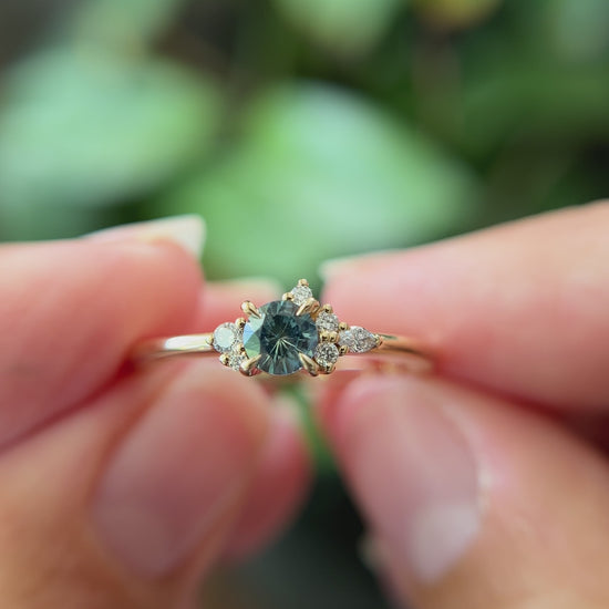 0.38ct round teal sapphire and natural diamond constellation engagement ring in 14k yellow gold, held in hands showing gemstone colour and faceting