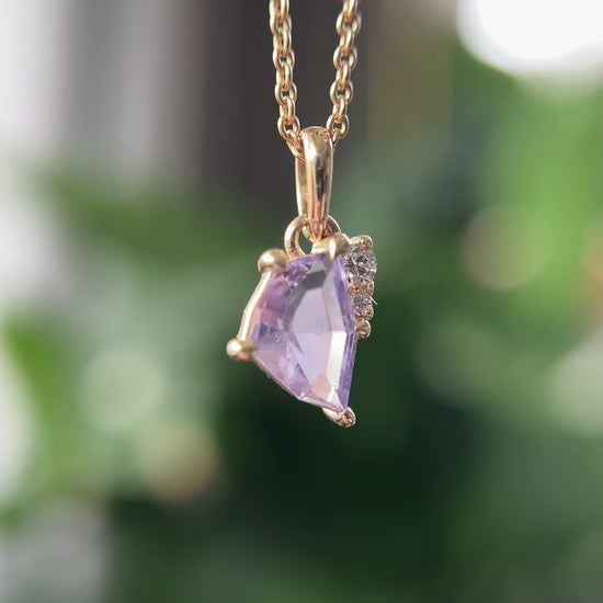 14k yellow gold pendant, set with a lavendar freeform sapphire and natural diamond accents. The pendant is hanging from a chain, in front of a green blurry background