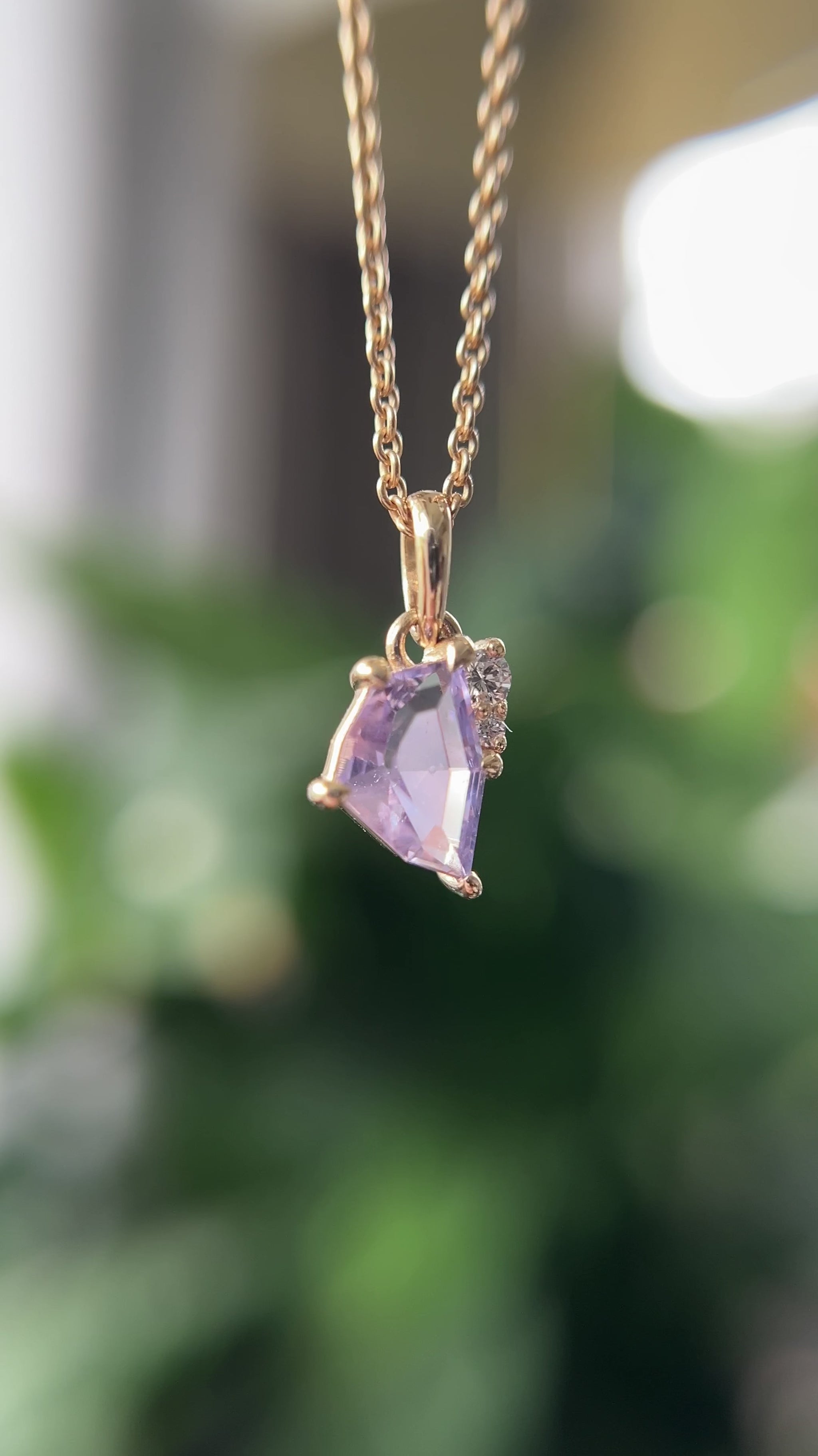 14k yellow gold pendant, set with a lavendar freeform sapphire and natural diamond accents. The pendant is hanging from a chain, in front of a green blurry background