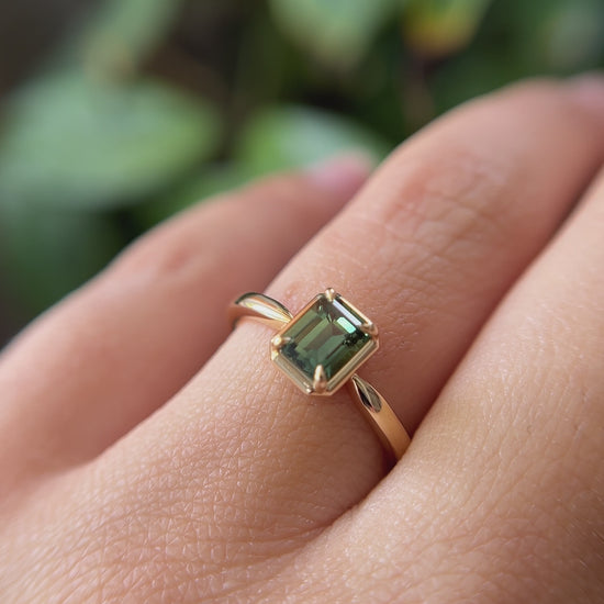 1.05ct emerald-cut forest green Madagascar sapphire solitaire engagement ring in 14k yellow gold, worn on finger showing colour and setting detail