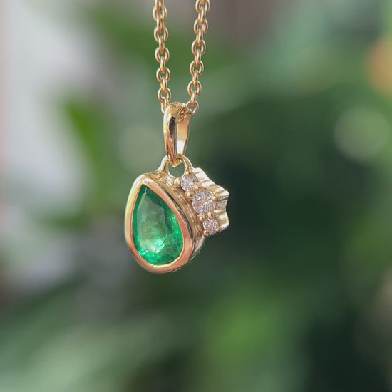 14k yellow gold pendant, set with a pear-shaped emerald and natural diamond accents. The pendant is hanging from a chain, in front of a green blurry background