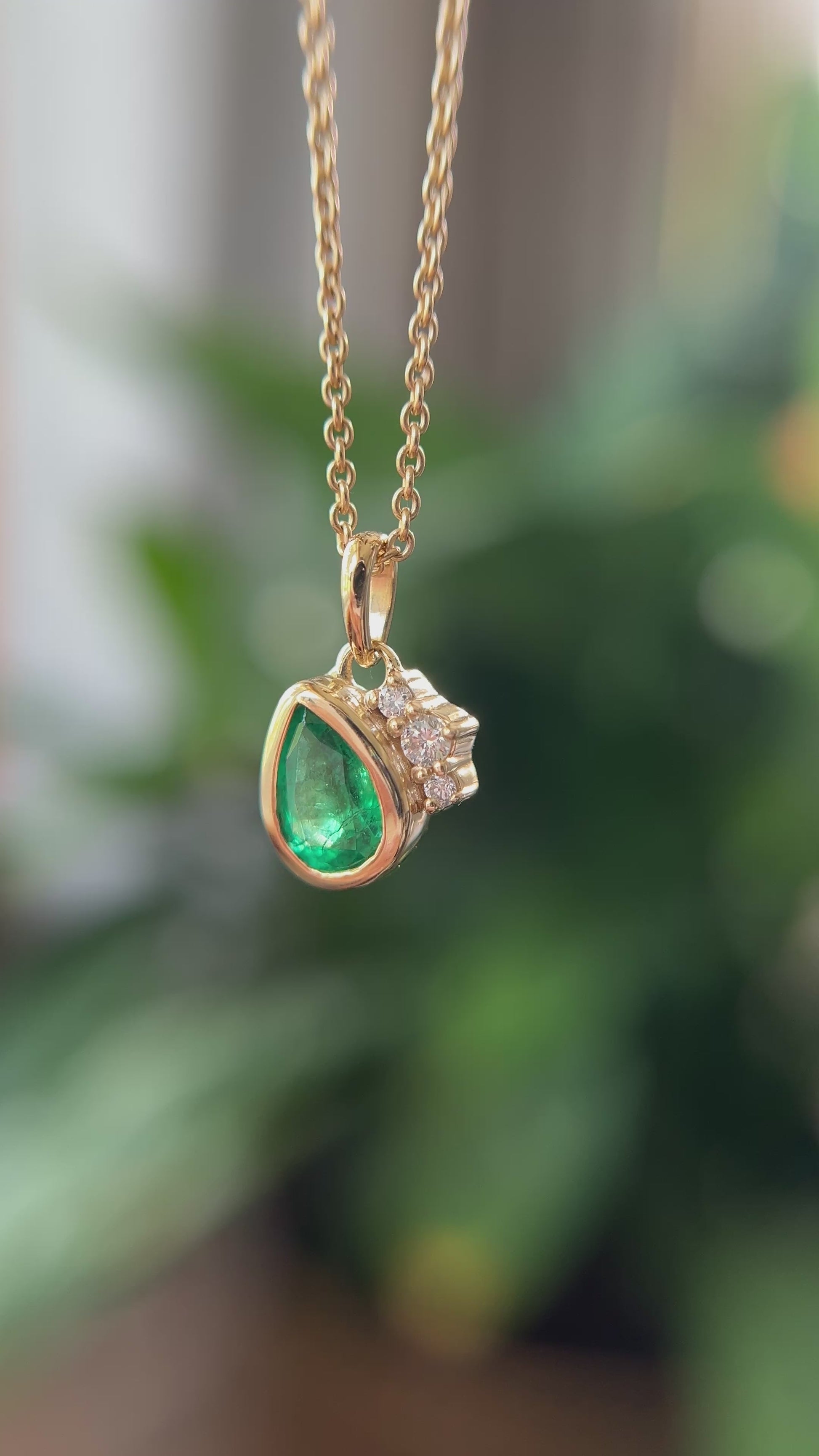 14k yellow gold pendant, set with a pear-shaped emerald and natural diamond accents. The pendant is hanging from a chain, in front of a green blurry background